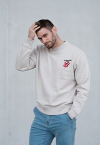 Sudadera beige con puños y dobladillo acanalados, con un logo bordado rojo de los Rolling Stones en el pecho izquierdo. Combina con jeans azul claro.