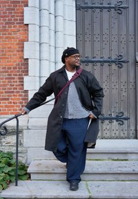 Manteau noir par-dessus un pull gris et une chemise blanche, pantalon gris large, chaussures noires et béret noir. Marches en pierre et porte en bois en arrière-plan.