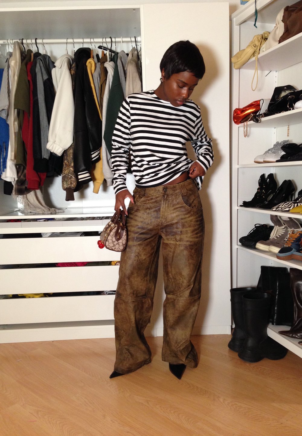 Femme portant un haut rayé à manches longues et un pantalon large marron, tenant un sac à main, debout dans une pièce avec un portant à vêtements et des étagères de chaussures et de sacs.