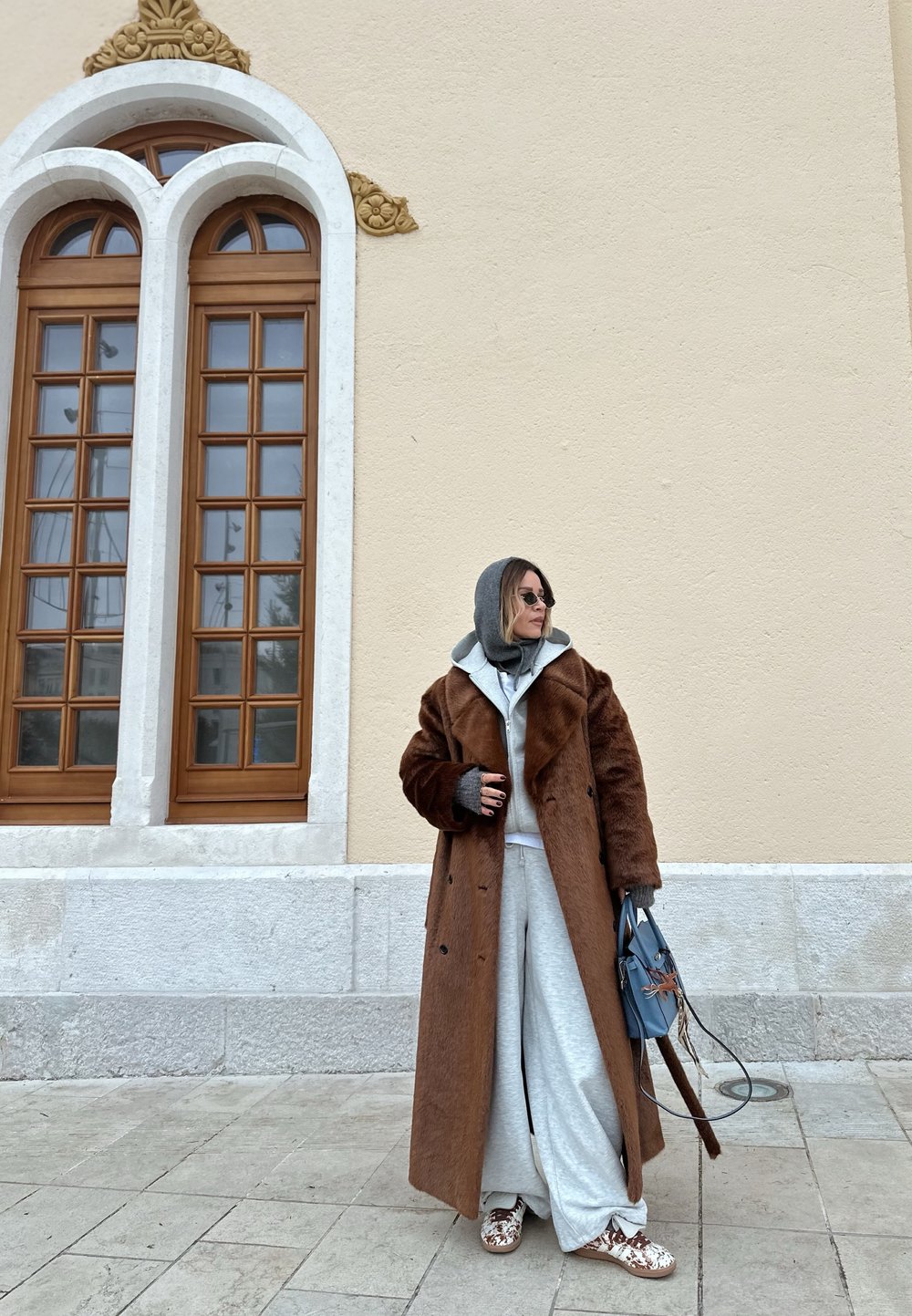 Personne portant un long manteau marron, un sweat à capuche gris, un pantalon large, des lunettes de soleil, et tenant un sac à main bleu, se tenant près d'un mur beige avec des fenêtres en arc.