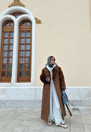 Persona vestida con un abrigo largo marrón, sudadera con capucha gris, pantalones anchos, gafas de sol y sosteniendo un bolso azul, de pie cerca de una pared beige con ventanas arqueadas.