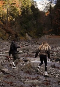 Dvě postavy procházejí přes kamenité koryto řeky, obklopené podzimním listím. Jedna má na sobě tmavou prošívanou bundu a zelené kalhoty; druhá hnědou prošívanou bundu a černé legíny.