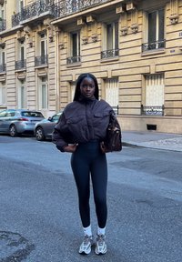 Kvinne iført en kort, mørkebrun jakke, svarte leggings, hvite sokker og sneakers står på en bygate mens hun holder en brun håndveske.