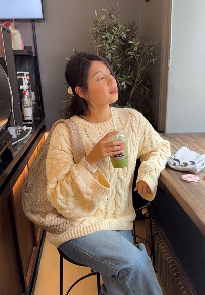 Suéter crema de punto trenzado, jeans azules, bolso trenzado beige, sosteniendo una bebida verde con una pajita, sentado en una mesa de madera en un ambiente de café.