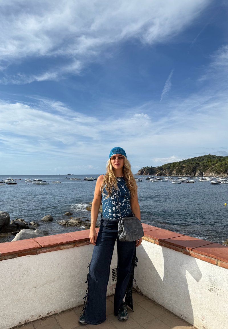 Top a canotta blu a fantasia, jeans larghi scuri con legami laterali, borsa a tracolla grigia e occhiali da sole. Sullo sfondo si vedono acqua e barche.