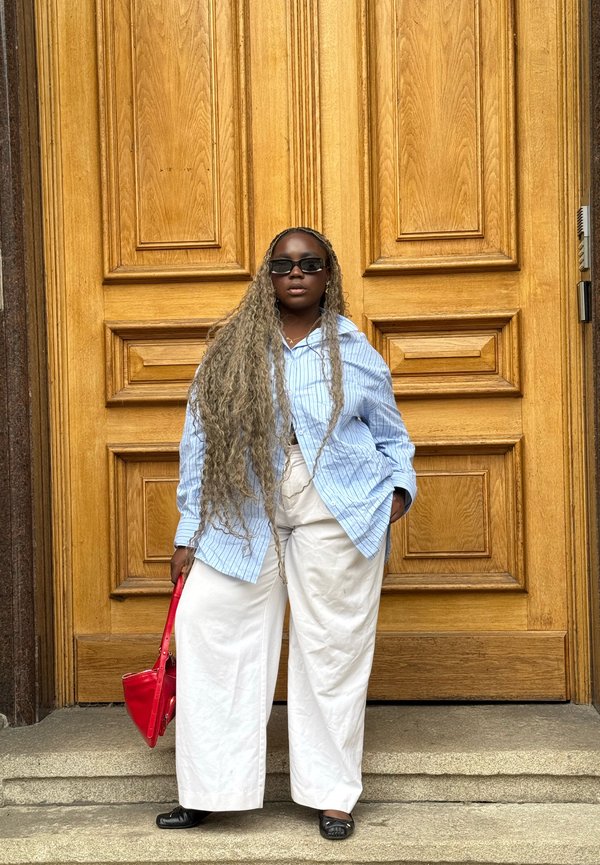 Femme aux longs cheveux bouclés portant des lunettes de soleil, une chemise à rayures bleues, un pantalon blanc, des chaussures noires, tenant un sac à main rouge, debout devant une grande porte en bois.