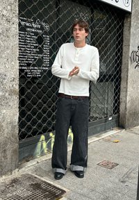 Chemise blanche à manches longues avec patte de boutonnage, associée à un jean noir à jambes larges avec ourlet effiloché. Chaussures noires. En arrière-plan, une devanture de magasin clôturée.