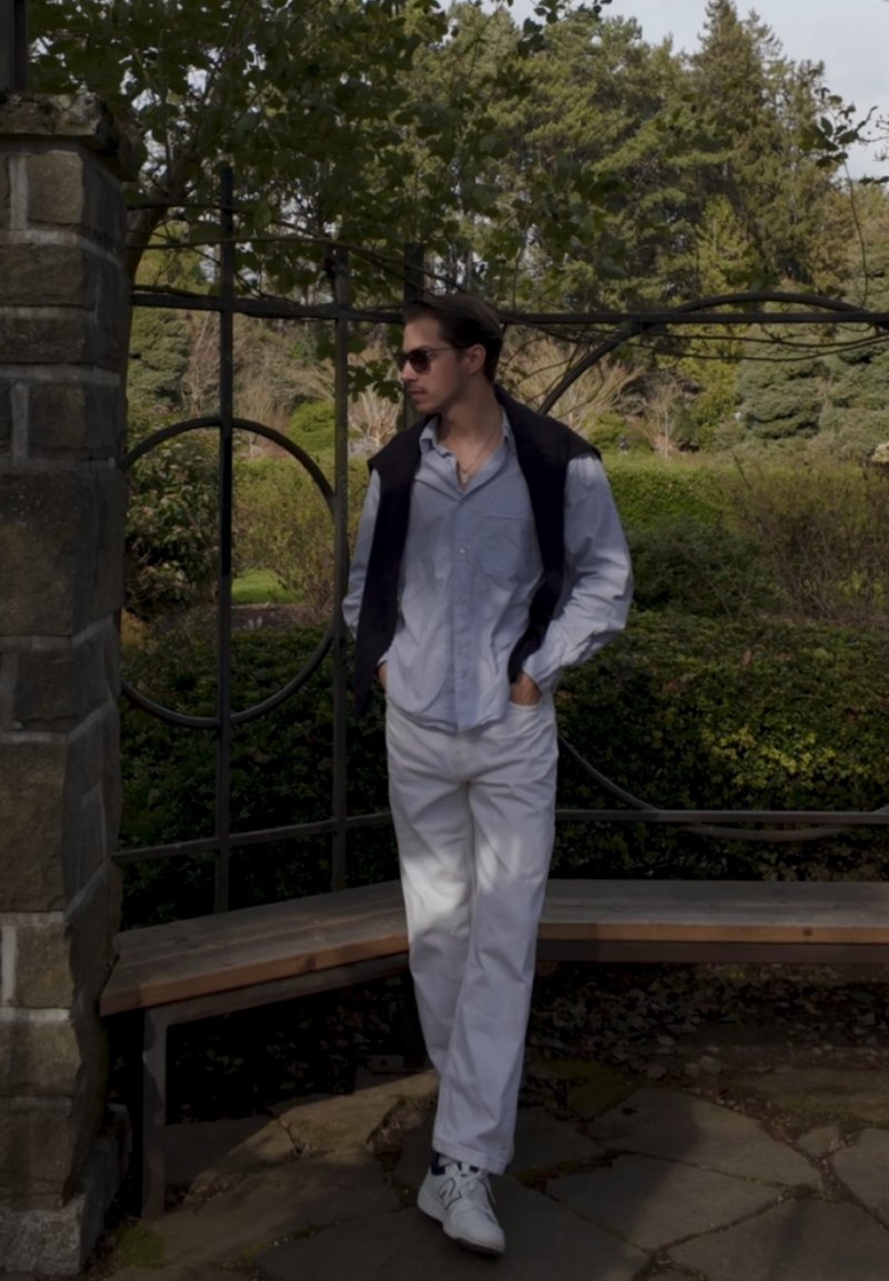 Mann mit Sonnenbrille, hellblauem Hemd, weißer Hose und Turnschuhen steht mit den Händen in den Taschen neben einer Steinsäule und einem Metall-Gartengitter.