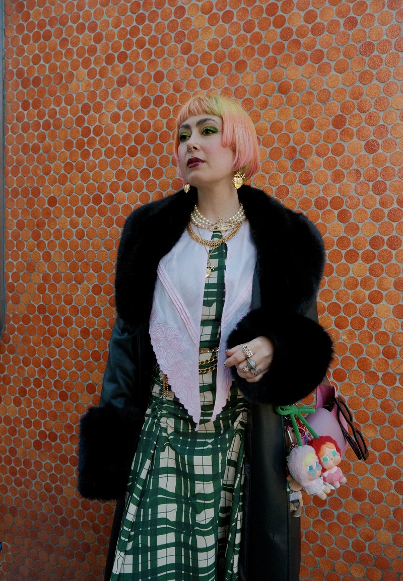 Stylish black coat with fur cuffs, layered over a green and white plaid dress. Pink silk scarf with embroidery; colorful accessories, including plush toys.