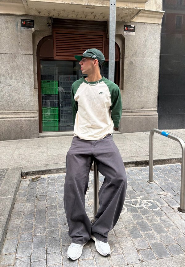 Jeune homme en t-shirt Nike vert et blanc, pantalon large gris et casquette verte appuyé contre un poteau métallique dans une rue pavée.