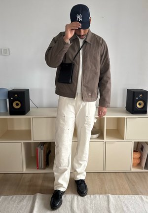 Chaqueta marrón, pantalones crema texturizados con acentos de costura, zapatos negros y una gorra azul marino. Un pequeño bolso negro cuelga del hombro.