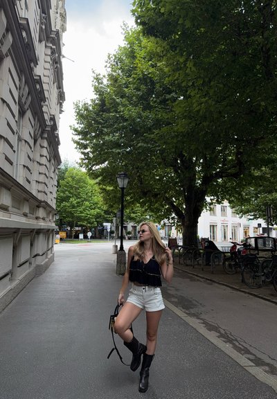 Top negro con volantes y ribetes dorados, pantalones cortos blancos y botas altas negras. De pie en una calle bordeada de árboles con edificios al fondo.