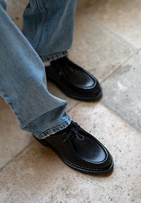 Scarpe di pelle nera con punta arrotondata, dettaglio cucito e lacci, abbinate a pantaloni in denim blu, seduto su un pavimento di pietra.