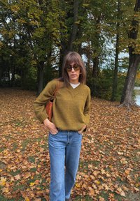 Oliivroheline V-kaelusega kampsun tekstuuriga viimistlusega, millele on sobitatud helesinised teksapüksid. Pruun punutud käekott. Taustal sügislilled.