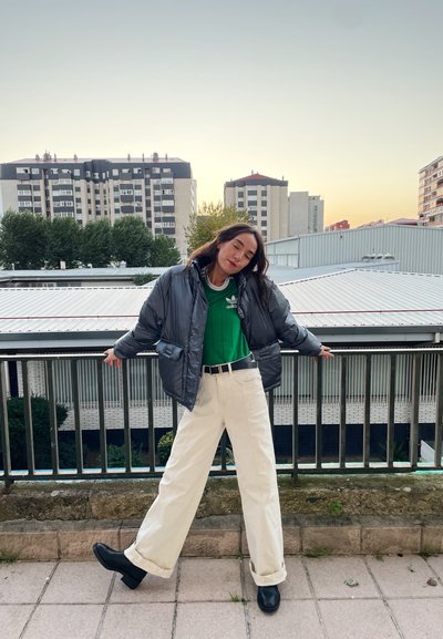 Chaqueta de plumas plateada, camiseta deportiva verde, pantalones anchos blancos, cinturón negro y botines tobilleros negros. Fondo sencillo con edificios urbanos.