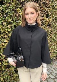 Cappotto corto nero con colletto alzato, dotato di maniche ampie. Accoppiato con un outfit beige e una piccola borsa nera con hardware dorato.
