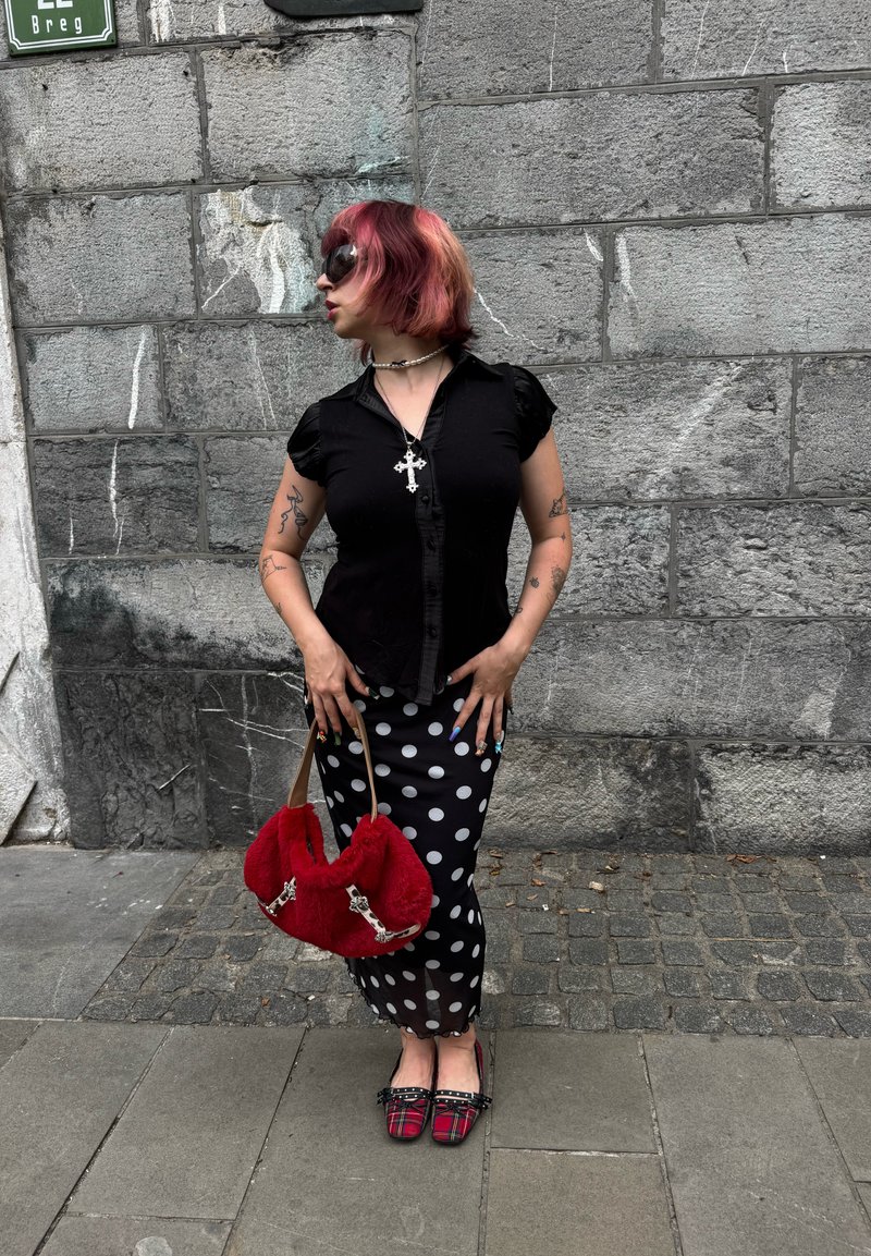 Personne avec de courts cheveux roses portant des lunettes de soleil, un haut noir, une jupe à pois, tenant un sac rouge, debout contre un mur en pierre gris sur le trottoir.