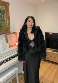 Femme portant une veste en fourrure noire et une robe en dentelle noire se tient à côté d'un piano blanc avec des partitions dans une pièce chaleureuse au sol en bois.