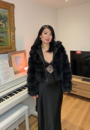 Mujer con chaqueta de piel negra y vestido de encaje negro de pie junto a un piano blanco con partituras en una habitación acogedora con suelo de madera.