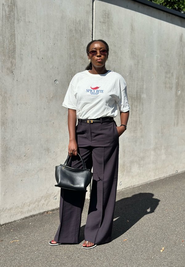Woman wearing sunglasses, white graphic t-shirt, black wide pants, and sandals, holding a black handbag, standing against a concrete wall outdoors.