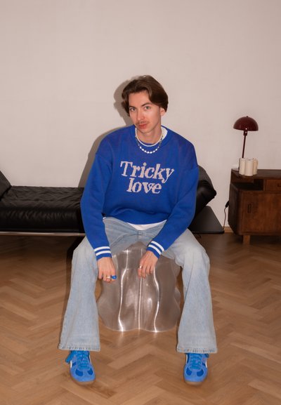 Persona joven con cabello castaño que lleva un suéter azul con la frase "Tricky love", jeans claros y zapatillas azules, sentada en una silla transparente en el interior.