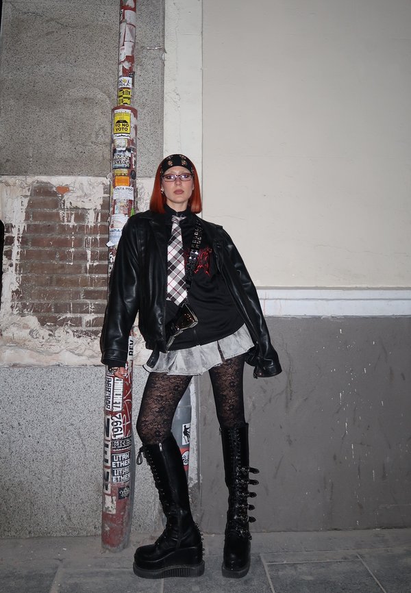 Chaqueta de cuero negra sobre una camisa negra, falda en capas, medias de encaje negro texturizadas y botas de caña alta con cordones. Los accesorios incluyen un pañuelo y gafas.