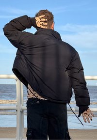 Doudoune noire avec col montant, tissu texturé et coupe oversize. Gants noirs avec un motif. En arrière-plan, de l'eau et une rambarde.