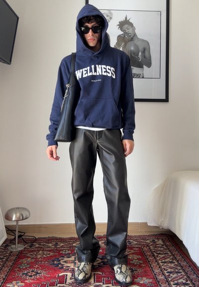 Sudadera navy con el texto "WELLNESS", pantalones de cuero sintético negros y botas de serpiente de estampado patrón. Accesorio: bolsa de hombro negra.