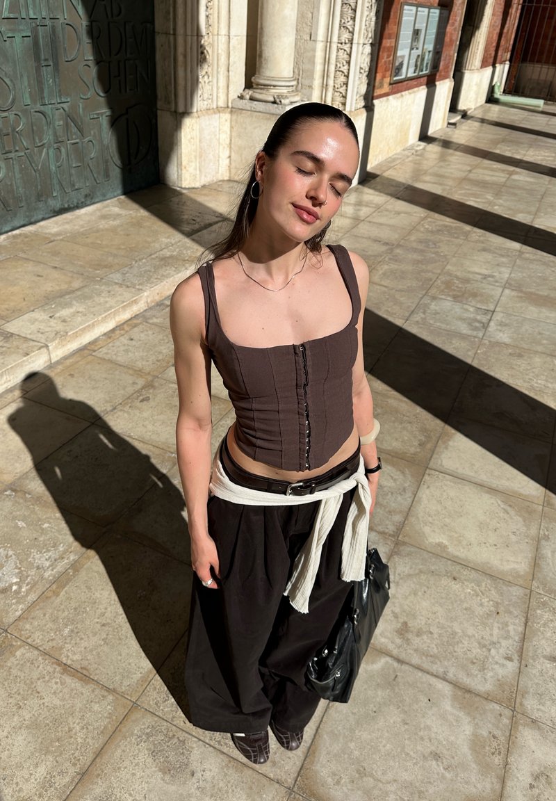 Donna in piedi su un percorso esterno piastrellato al sole con gli occhi chiusi, indossa un top scuro, pantaloni larghi, orecchini e tiene una borsa nera.