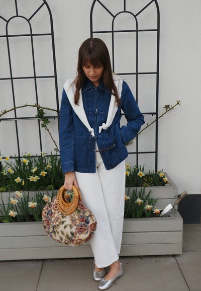 Mujer con chaqueta vaquera y pantalones blancos sosteniendo un bolso con estampado floral, de pie junto a macetas con narcisos en flor contra una pared blanca.