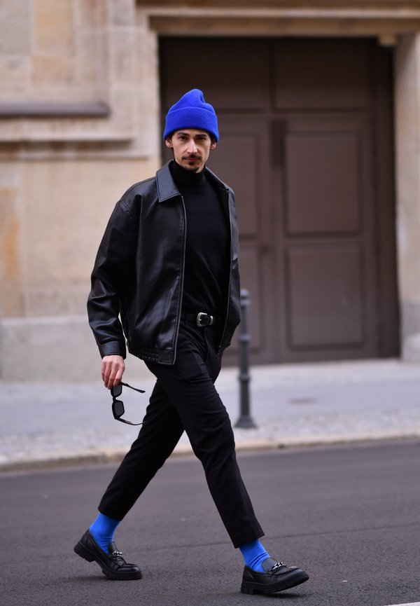 Chaqueta de cuero negra, suéter de cuello alto negro, pantalones cortos negros, calcetines azules, zapatos negros con detalles, y un gorro azul; sosteniendo unas gafas de sol.