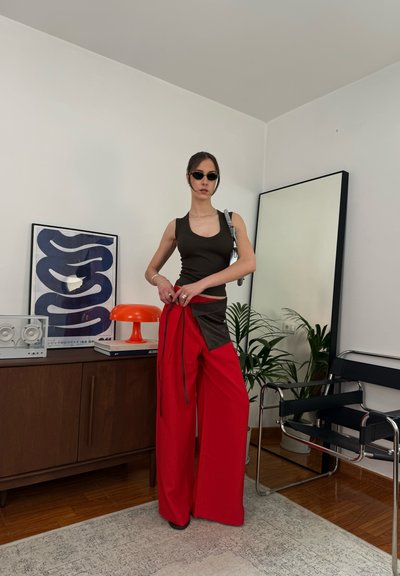 Mujer con top negro sin mangas, pantalones anchos rojos y gafas de sol, está en una sala de estar moderna con espejo, plantas y muebles de mediados de siglo.