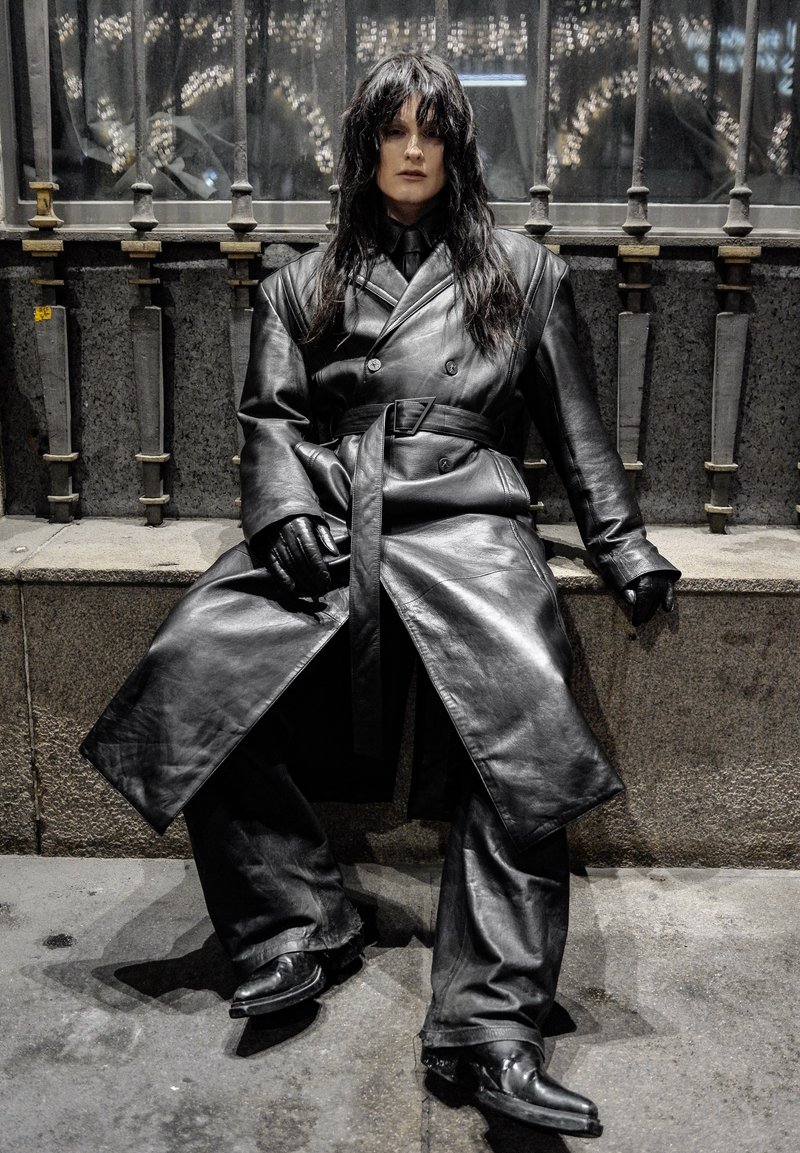 Trench-coat en cuir noir avec ceinture, assorti à des gants et un pantalon en cuir. Design épuré, col montant et bottes pointues.