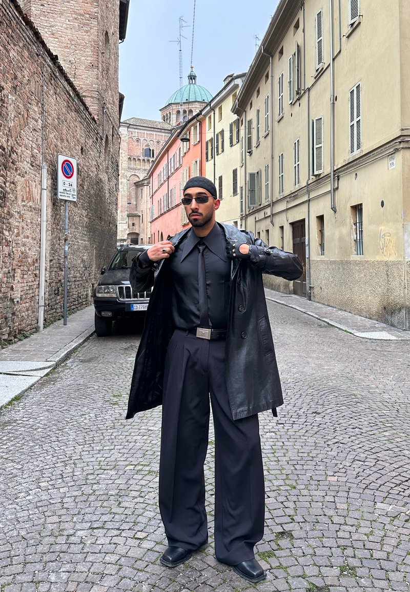 Mann in schwarzem Lederjacke, weiten Hosen und Sonnenbrille steht auf einer Kopfsteinpflasterstraße mit historischen Gebäuden und geparktem Auto im Hintergrund.