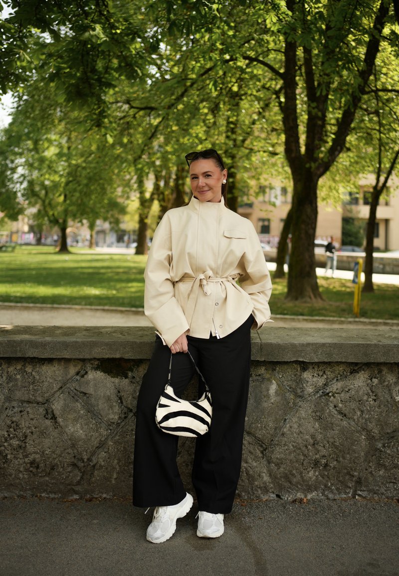 Femme en veste beige oversize et pantalon noir, debout contre un mur de pierre, tenant un sac à motif zèbre, avec des arbres et un parc en arrière-plan.