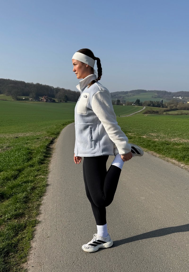 Femeie îmbrăcată în haine sportive, întinzând cvadricepsul pe un drum de țară cu câmpuri verzi și dealuri sub un cer senin.