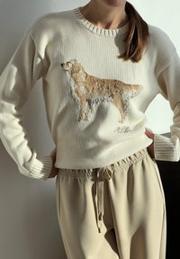 Krémový pletený sveter s vyšívaným dizajnom psa v béžovej a hnedej farbe, s rebrovaným golierom a manžetami. Spolu s svetlo béžovými nohavicami.