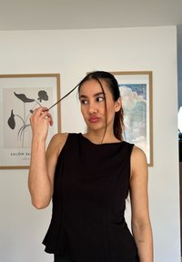 Čierny top bez rukávov s vysokým výstrihom, hladkou textúrou a priliehavým strihom. Modelka drži strandu vlasov pri svetlej stene.