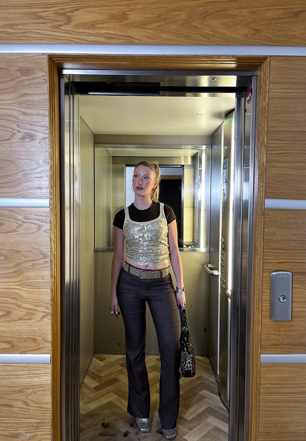 Ung kvinde står inde i en moderne elevator med åbne døre, iført en palietteret top, mørke bukser og holder en leopardmønstret taske.