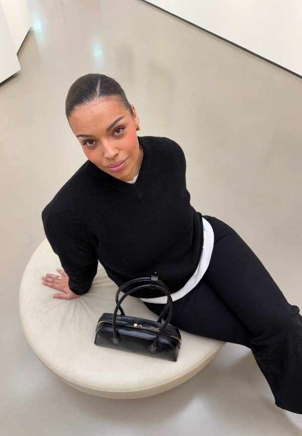 Vrouw met donker haar, gekleed in een zwarte trui en broek, zittend op een ronde beige stoel met een zwarte handtas naast haar op een gladde vloer