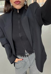 Fekete gyapjú zakó struktúrált vállakkal egy bordázott fekete felső fölött, elöl cipzárral. A szettet szürke szabott nadrág és szegecselt öv egészíti ki.