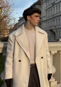 Manteau long en mélange de laine blanche avec grands revers et boutons noirs, porté sur un pull en maille beige clair et un pantalon foncé, avec des gants en cuir.