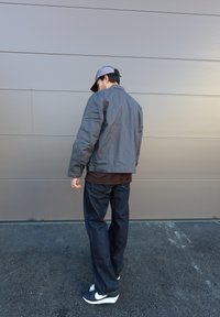 Chaqueta gris con cuello abotonado, camisa marrón debajo, jeans azul oscuro y zapatillas Nike negras, vista desde atrás.