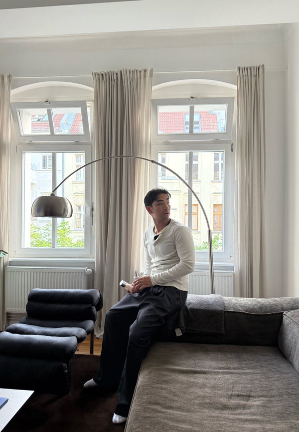 Mand, der sidder på kanten af en grå sofa og holder en fjernbetjening, kigger til siden i en lys stue med store vinduer og gardiner.
