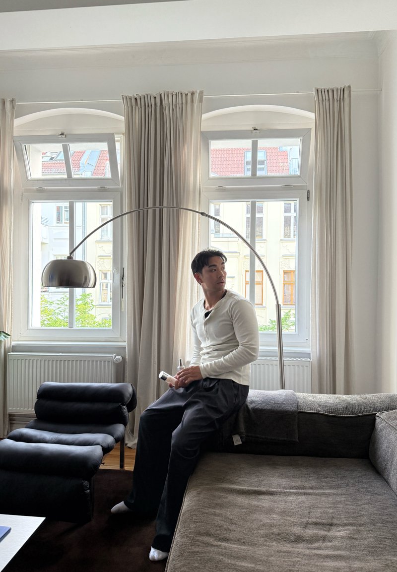 Homme assis au bord d’un canapé gris tenant une télécommande, regardant sur le côté dans un salon lumineux avec de grandes fenêtres et des rideaux.