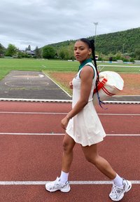 Robe de sport blanche avec col vert, associée à des chaussures New Balance blanches et un sac à dos crème avec des accents orange, marchant sur une piste.