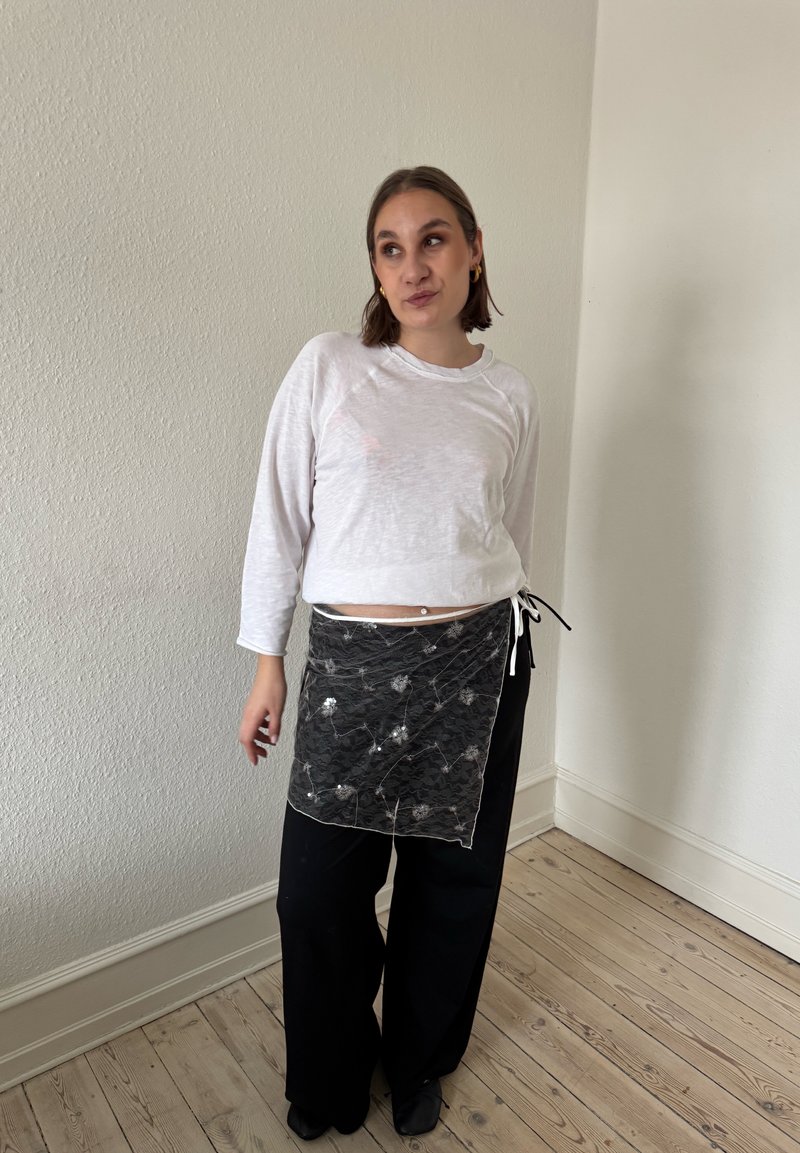 Femme aux cheveux courts bruns portant un haut blanc à manches longues, un pantalon noir à jambes larges et un tablier en dentelle noir, debout à l'intérieur sur un sol en bois.