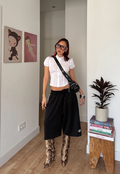 Mujer con blusa blanca de mangas abullonadas, pantalones cortos negros de pierna ancha, botas con estampado de serpiente, bolso cruzado negro y gafas de gran tamaño, de pie en una habitación moderna.