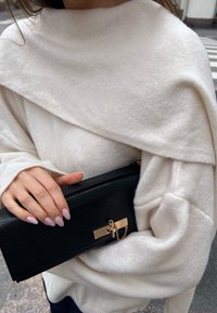 Pulôver branco oversized com um gola alta; clutch preta texturizada com detalhes em ouro e corrente segurada numa mão com unhas rosa claro.