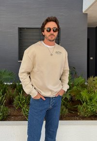 Beżowa bluza z długim rękawem, z logo z przodu. Połączona z niebieskimi dżinsami, na tle zielonego listowie i szarej ściany.